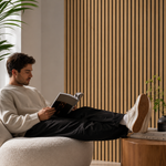 A man relaxes in a cream boucle lounge chair, reading a book with his legs stretched, set against a modern background with a slatted wood wall and a side table.